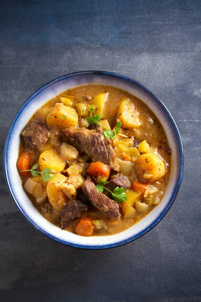 Beef meat stewed with potatoes, carrots and spices in bowl on dark gray background. Overhead vertical image