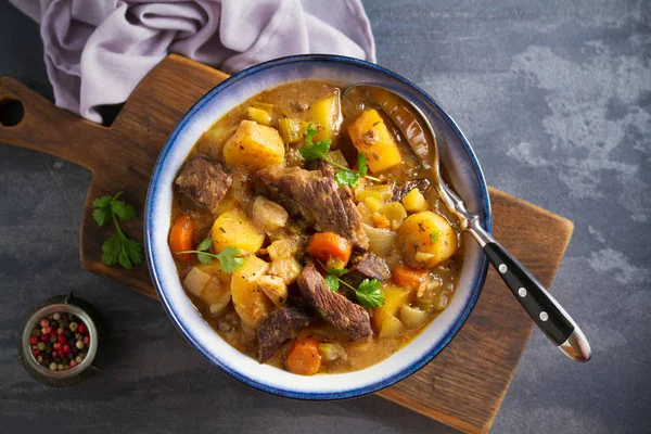 Beef meat stewed with potatoes, carrots and spices in bowl on dark gray background. Overhead horizontal image