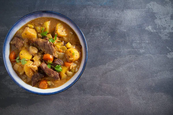 Beef meat stewed with potatoes, carrots and spices in bowl on dark gray background. Overhead horizontal image, room for text