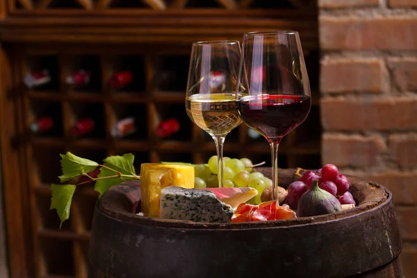 ワインセラーの赤と白のワインのグラスには ワイン樽のブドウ チーズ プロシュット イチジク ナッツが添えられています — ストック写真
