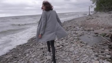 Gri montlu bir kız deniz kıyısındaki taştan bir sahil boyunca yürüyor, saçları güçlü bir rüzgardan gelişiyor, deniz fırtınaları...