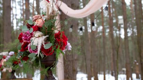 露に赤いバラで構成された結婚式の花束は冬の松林に電球で濡れたアーチに対して花瓶に立っています — ストック動画