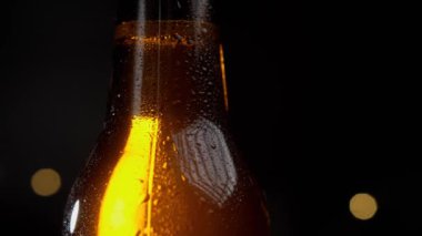 Su damlaları yavaş yavaş soğuk bir bira şişesine akar. Siyah arka planda şişe altın renginde parlar.