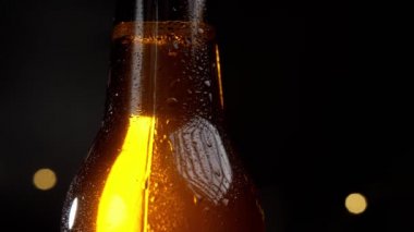 Su damlaları yavaş yavaş soğuk bir bira şişesine akar. Siyah arka planda şişe altın renginde parlar.