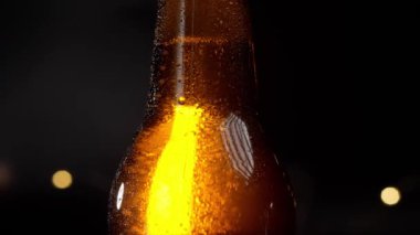 Damlalarla kaplı soğuk bir bira şişesi. Siyah arka planda makro çekim. Şişe altın gibi parlıyor, damlalar yavaşça aşağı akıyor.