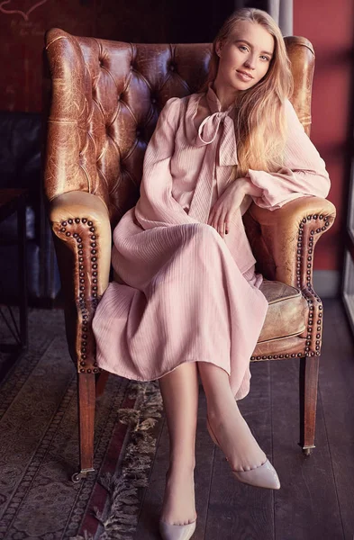 Beautiful elegant blonde woman in pink dress sitting in leather armchair — Stock Photo, Image