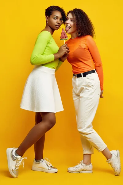 Két fiatal, gyönyörű hipster lány színes nyári ruhában nyalókával. — Stock Fotó