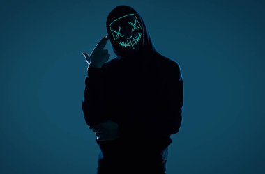 Siyah kapüşonlu isimsiz bir adam yüzünü neon bir maskenin arkasına gizliyor.