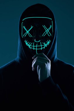 Siyah kapüşonlu isimsiz bir adam yüzünü neon bir maskenin arkasına gizliyor.