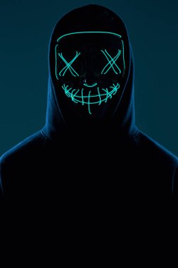 Siyah kapüşonlu isimsiz bir adam yüzünü neon bir maskenin arkasına gizliyor.