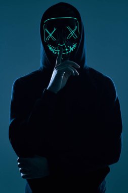 Siyah kapüşonlu isimsiz bir adam yüzünü neon bir maskenin arkasına gizliyor.