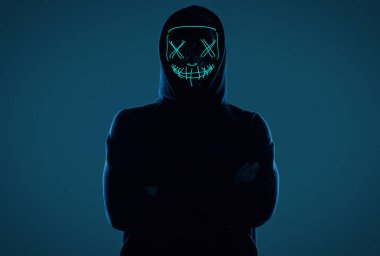 Siyah kapüşonlu isimsiz bir adam yüzünü neon bir maskenin arkasına gizliyor.