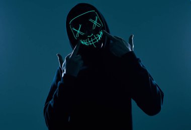 Siyah kapüşonlu isimsiz bir adam yüzünü neon bir maskenin arkasına gizliyor.