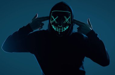 Siyah kapüşonlu isimsiz bir adam yüzünü neon bir maskenin arkasına gizliyor.