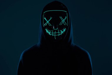 Siyah kapüşonlu isimsiz bir adam yüzünü neon bir maskenin arkasına gizliyor.