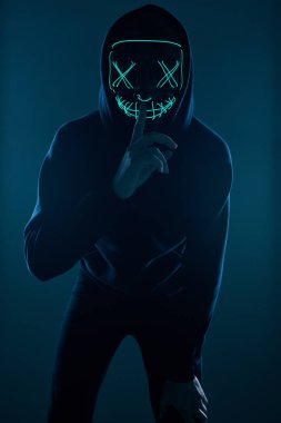 Siyah kapüşonlu isimsiz bir adam yüzünü neon bir maskenin arkasına gizliyor.