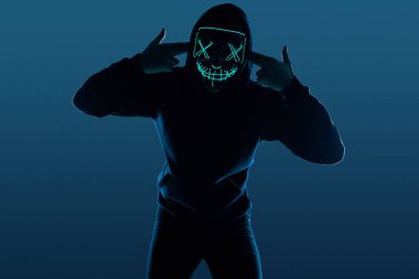 Siyah kapüşonlu isimsiz bir adam yüzünü neon bir maskenin arkasına gizliyor.