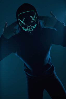 Siyah kapüşonlu isimsiz bir adam yüzünü neon bir maskenin arkasına gizliyor.