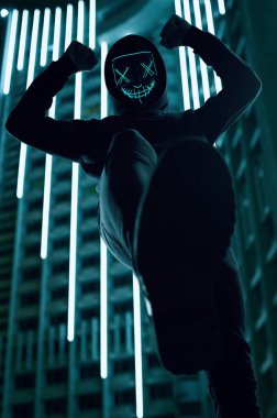 Siyah kapüşonlu isimsiz bir adam yüzünü neon bir maskenin arkasına gizliyor.
