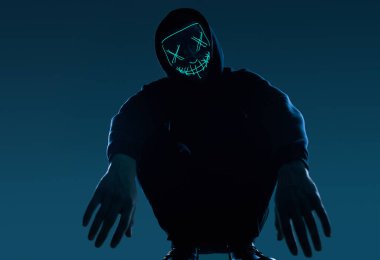 Siyah kapüşonlu isimsiz bir adam yüzünü neon bir maskenin arkasına gizliyor.