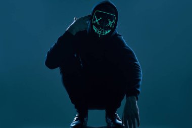 Siyah kapüşonlu isimsiz bir adam yüzünü neon bir maskenin arkasına gizliyor.