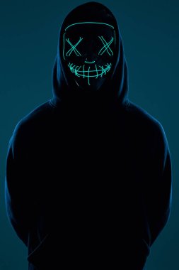 Siyah kapüşonlu isimsiz bir adam yüzünü neon bir maskenin arkasına gizliyor.