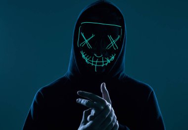 Siyah kapüşonlu isimsiz bir adam yüzünü neon bir maskenin arkasına gizliyor.