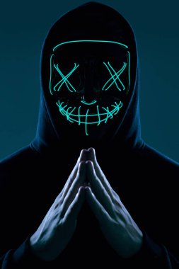 Siyah kapüşonlu isimsiz bir adam yüzünü neon bir maskenin arkasına gizliyor.