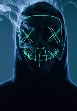 İsimsiz bir adam renkli bir dumanın içinde yüzünü neon maskenin arkasına gizliyor.