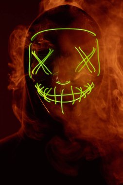 İsimsiz bir adam renkli bir dumanın içinde yüzünü neon maskenin arkasına gizliyor.