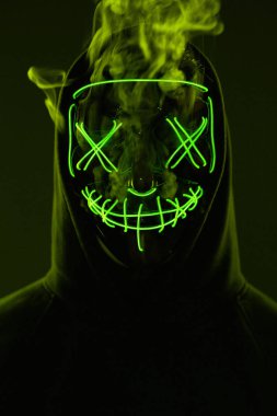 İsimsiz bir adam renkli bir dumanın içinde yüzünü neon maskenin arkasına gizliyor.