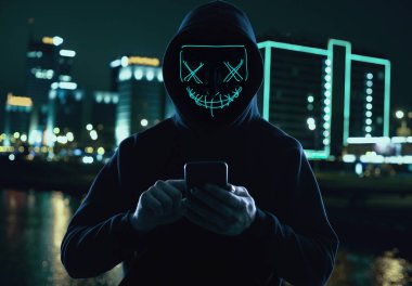 Anonymous man in a black hoodie and neon mask hacking into a smartphone clipart