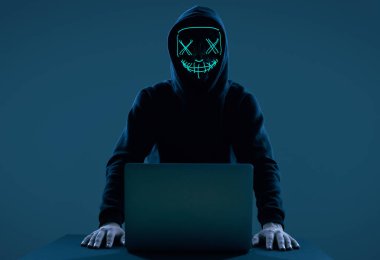 Anonymous man in a black hoodie and neon mask hacking into a computer clipart