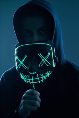 Siyah kapüşonlu isimsiz bir adam yüzünü neon bir maskenin arkasına gizliyor.