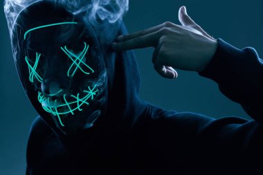 Siyah kapüşonlu isimsiz bir adam yüzünü neon bir maskenin arkasına gizliyor.