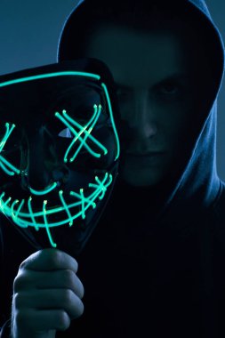 Siyah kapüşonlu isimsiz bir adam yüzünü neon bir maskenin arkasına gizliyor.
