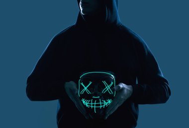 Siyah kapüşonlu isimsiz bir adam yüzünü neon bir maskenin arkasına gizliyor.
