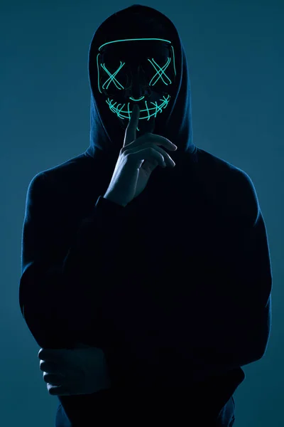 Siyah kapüşonlu isimsiz bir adam yüzünü neon bir maskenin arkasına gizliyor.