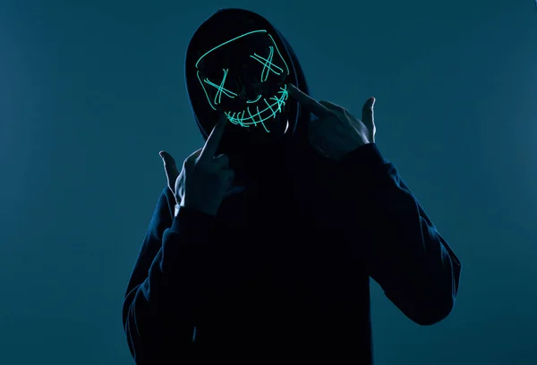 Siyah kapüşonlu isimsiz bir adam yüzünü neon bir maskenin arkasına gizliyor.