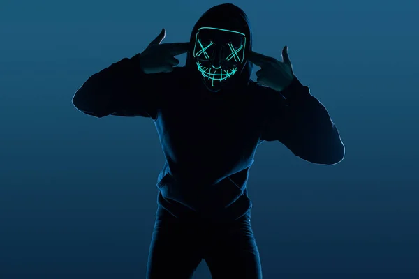 Névtelen férfi fekete kapucnis pulcsiban, neon maszk mögé rejtette az arcát. — Stock Fotó