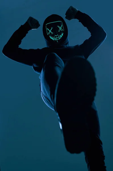 Hombre anónimo con capucha negra escondiendo su cara detrás de una máscara de neón — Foto de Stock