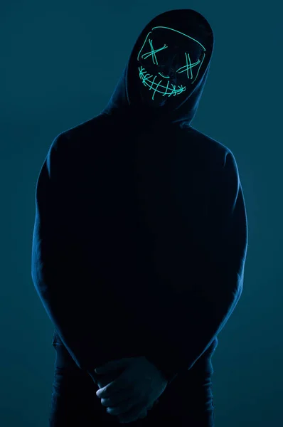 Siyah kapüşonlu isimsiz bir adam yüzünü neon bir maskenin arkasına gizliyor. — Stok fotoğraf
