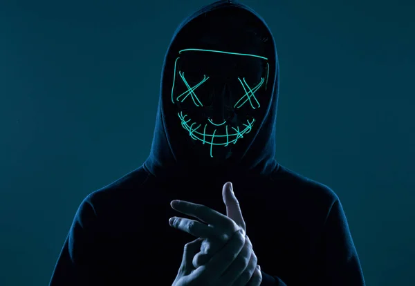 Siyah kapüşonlu isimsiz bir adam yüzünü neon bir maskenin arkasına gizliyor.