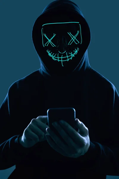 Siyah kapüşonlu ve neon maskeli isimsiz bir adam akıllı telefonu hackliyor. — Stok fotoğraf