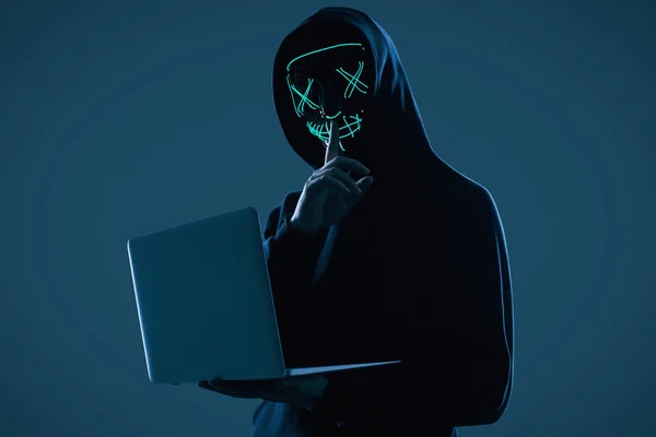 Анонимный человек в черной толстовке и неоновой маске, взламывающий компьютер — стоковое фото
