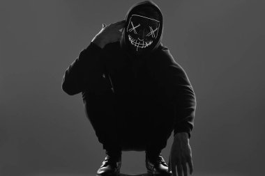 Siyah kapüşonlu isimsiz bir adam yüzünü neon bir maskenin arkasına gizliyor.