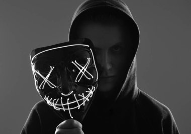 Siyah kapüşonlu isimsiz bir adam yüzünü neon bir maskenin arkasına gizliyor.