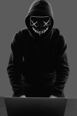 Anonymous man in a black hoodie and neon mask hacking into a com clipart