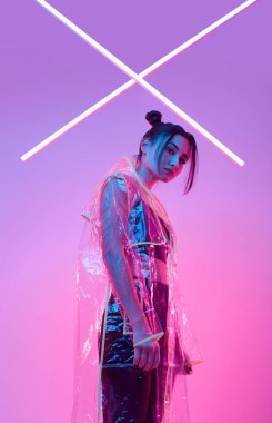 Renkli parlak neon ışıkların etrafında şık bir yağmurlukla stüdyoda poz veren zarif Asyalı bir kadının portresi.