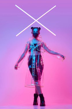 Güzel virüs maskeli Asyalı kadın portresi yüz koruması ve renkli parlak uv ışıkları etrafında yağmurluk stüdyoda poz veriyor.